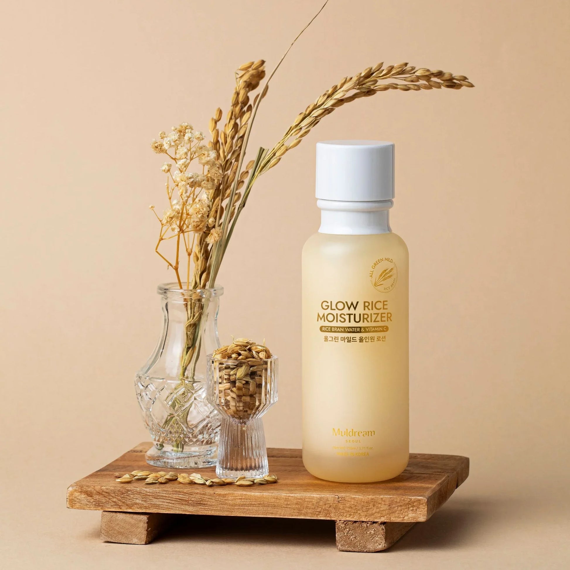 Glow Rice Moisturizer with rice bran water and vitamin C, displayed with dried rice stalks and grains on a wooden tray